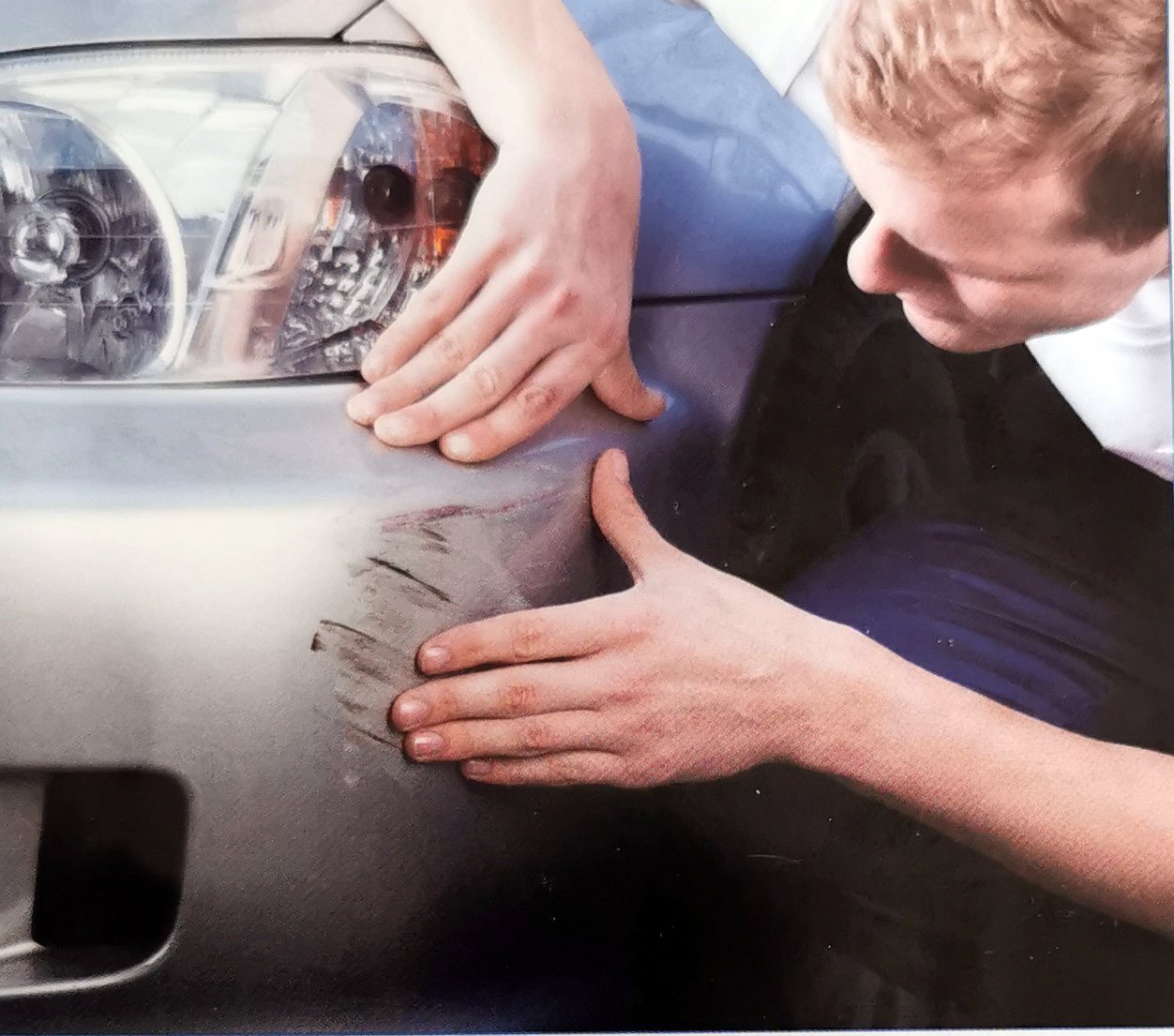 Hände prüfen einen Kratzer an der Stoßstange eines silbernen Autos
